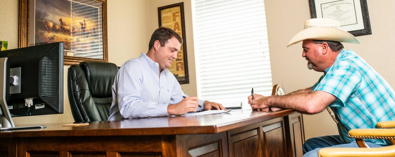 Two men signing documents in an office, one is sitting behind the desk with a computer on his side and the other is sitting in front wearing a hat.