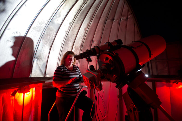 A woman observes the night sky using a telescope inside a dome observatory illuminated by red lighting.