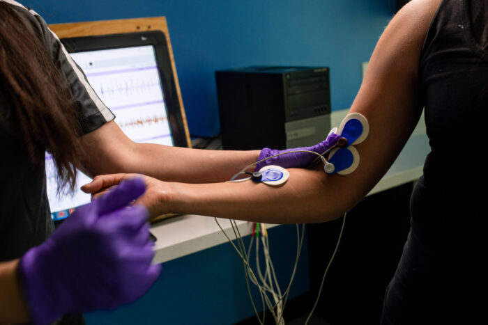 A person wearing purple gloves attaches electrodes to another person's arm while monitoring data on a computer screen in a medical or research setting.
