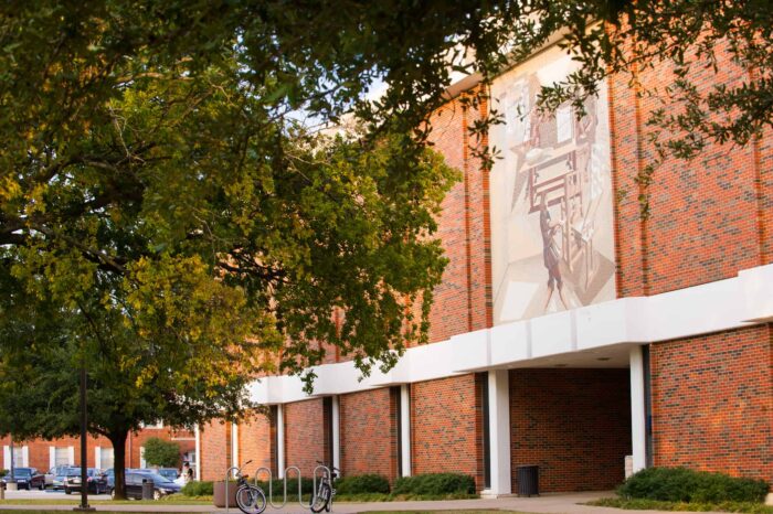 Brick building with large mural visible, framed by leafy green trees. White architectural details contrast with red bricks. Sunlight casts warm glow on facade. Campus or institutional setting.