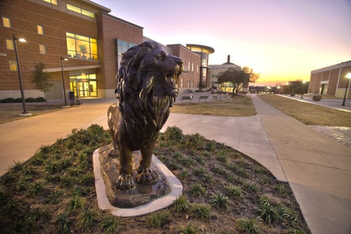 Lion statue in front of a bricked building. It is in the evening and the focus is on the lion.