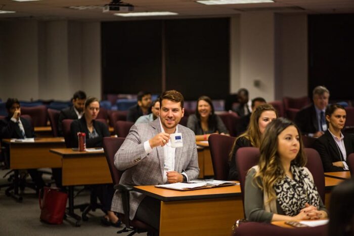 A group of people sit in a classroom with desks arranged in rows during a formal event. A man in a gray suit holds up a coffee cup while smiling. Other attendees are engaged and attentively looking towards the front.