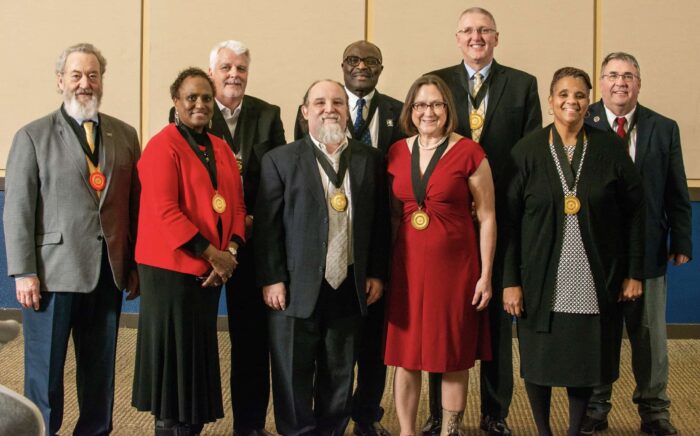 A group of nine adults, dressed in formal attire, stands together smiling for a photo. Some are wearing medallions around their necks. The background contains beige paneling. The group includes men and women, with a mix of ages and ethnicities.