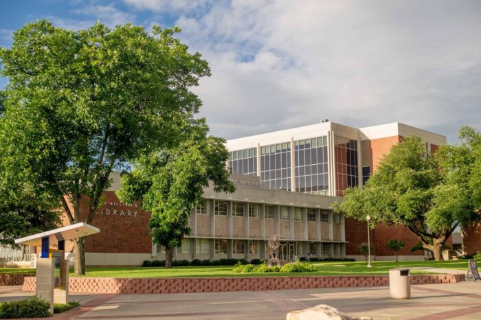 Modern university library building with glass facade and brick exterior. Cloudy sky background. There are a few trees in front of the building and a "lion" Sign reads "Gee Gillan" on the side