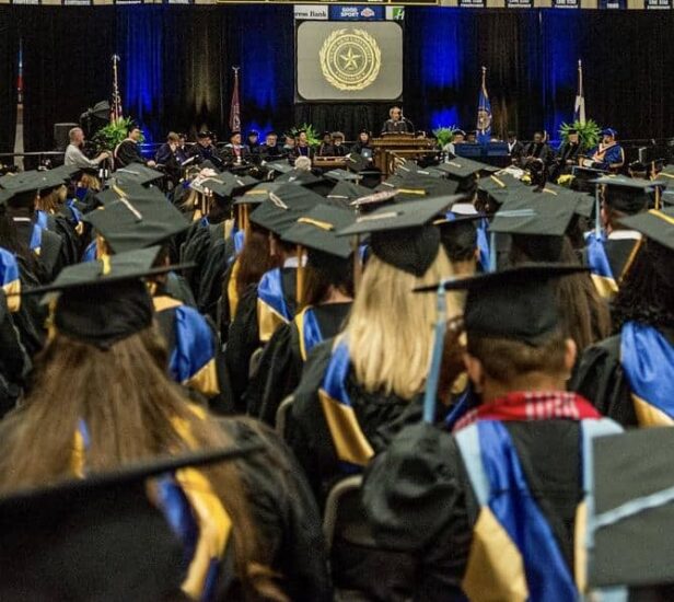 A large group of graduates in caps and gowns sit facing a stage during a commencement ceremony. The stage has officials and a podium, with a projection screen displaying a seal in the background. The venue is decorated with flags and banners.