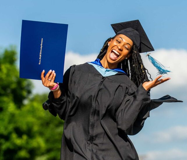 A joyful graduate in a black cap and gown smiles widely while holding up a blue diploma cover in one hand and gesturing with the other. The background features a clear sky and greenery.