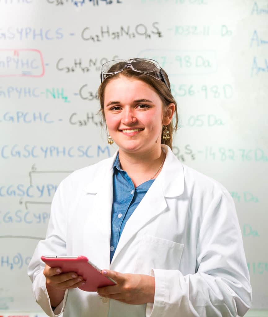 A student in a lab coat in front of a white board filled with chemistry codes.
