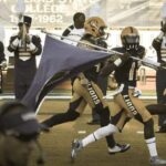 a group of football player running to the field with a flag.