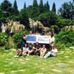 A group of students in front of a garden area holding the university sign.