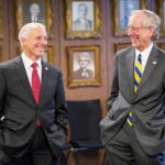 Two individual wearing business suits have their hands in their pocket and smiling randomly. They are in front of a wall with pictures.