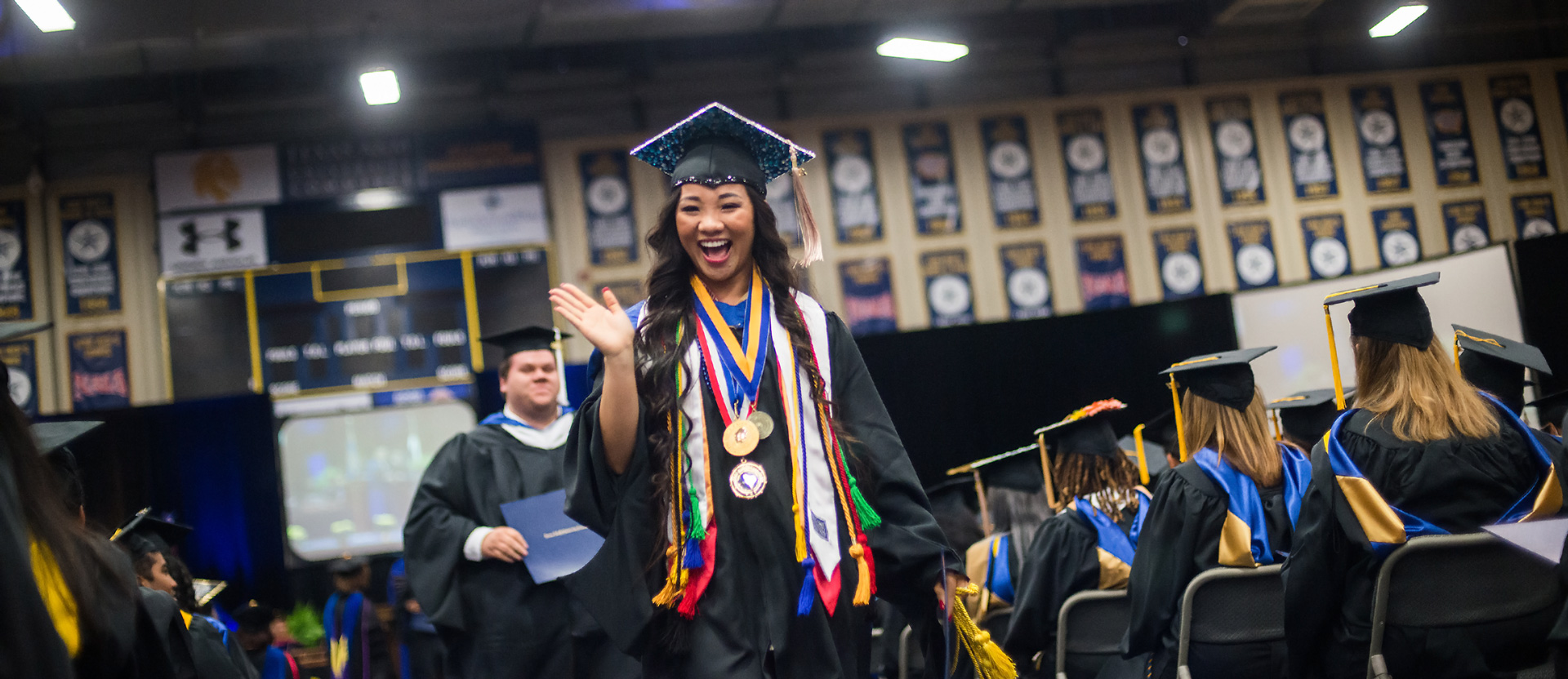 woman wearing medals and graduation gown waving during graduation ceremony