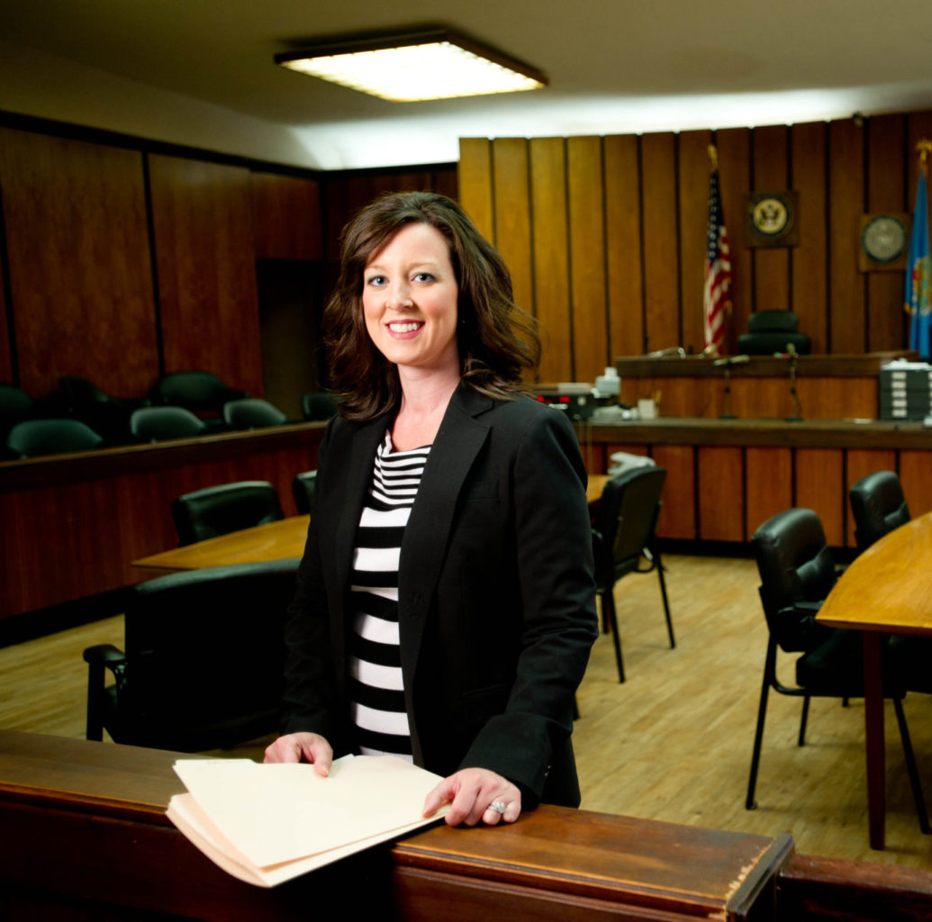 A female dressed professionally standing in a court room smiling at the camera.