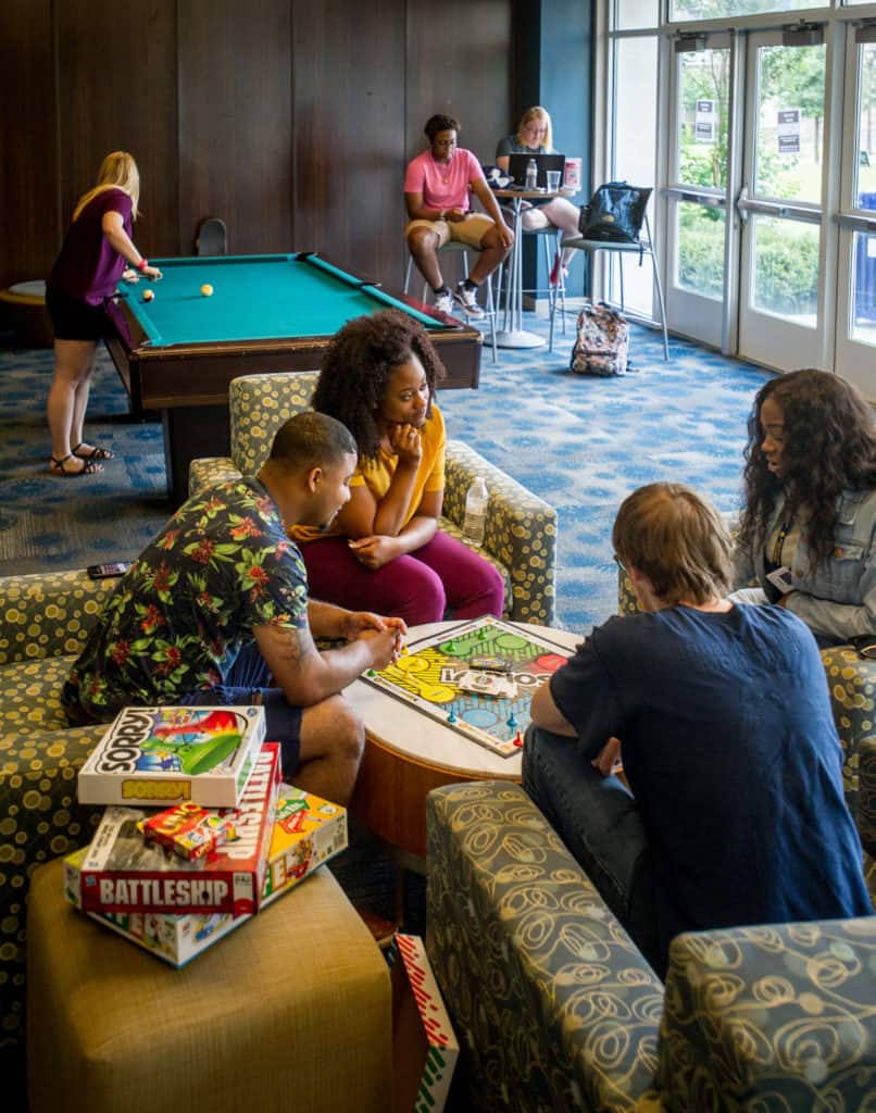 A group of student in a room speaking and playing pool.