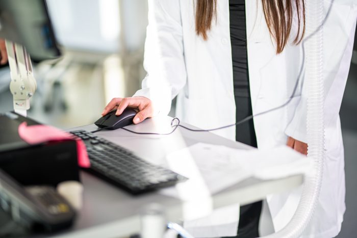 A person in a lab coat operates a computer mouse while standing at a workstation, with a keyboard and monitor visible on the desk.