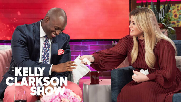 A man in a suit and tie is laughing while a woman in a red dress hands him a tissue. They are seated on a talk show set with a pink and purple background. The man looks joyful, and the woman is smiling warmly. Text on the image reads "The Kelly Clarkson Show.