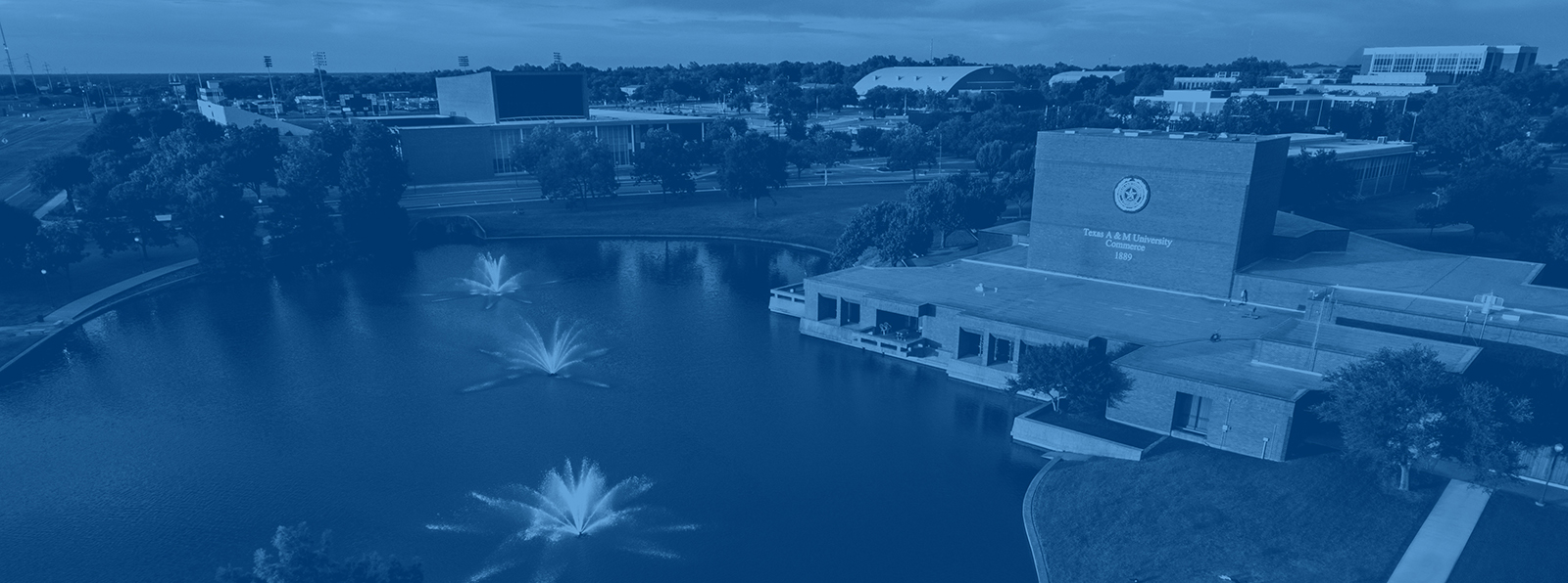 Aerial view of East Texas A&M University campus featuring a peaceful pond with three fountains, surrounding trees, and campus buildings in the background.