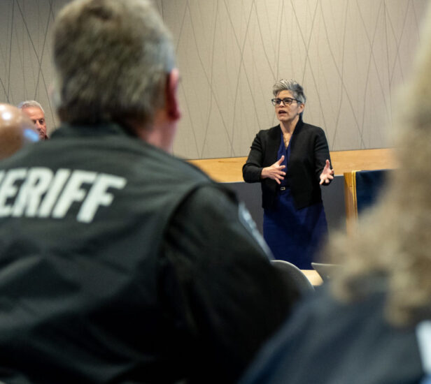 A woman wearing glasses and a dark blazer speaks in front of a group, gesturing with her hands. In the forefront, a man with "Sheriff" on his jacket listens attentively.