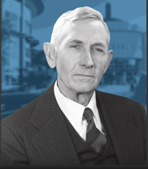 Black-and-white portrait of an elderly man with short, neatly combed hair, wearing a three-piece suit with a striped tie. He has a serious expression. The background features a blue-toned image of large, abstract buildings.