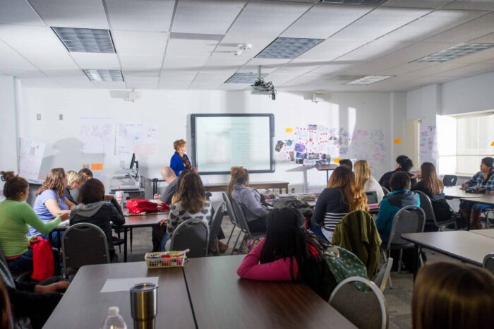 A classroom with a teacher standing at the front near a projected presentation. Students sit at tables, attentively facing the teacher, with posters on the walls.