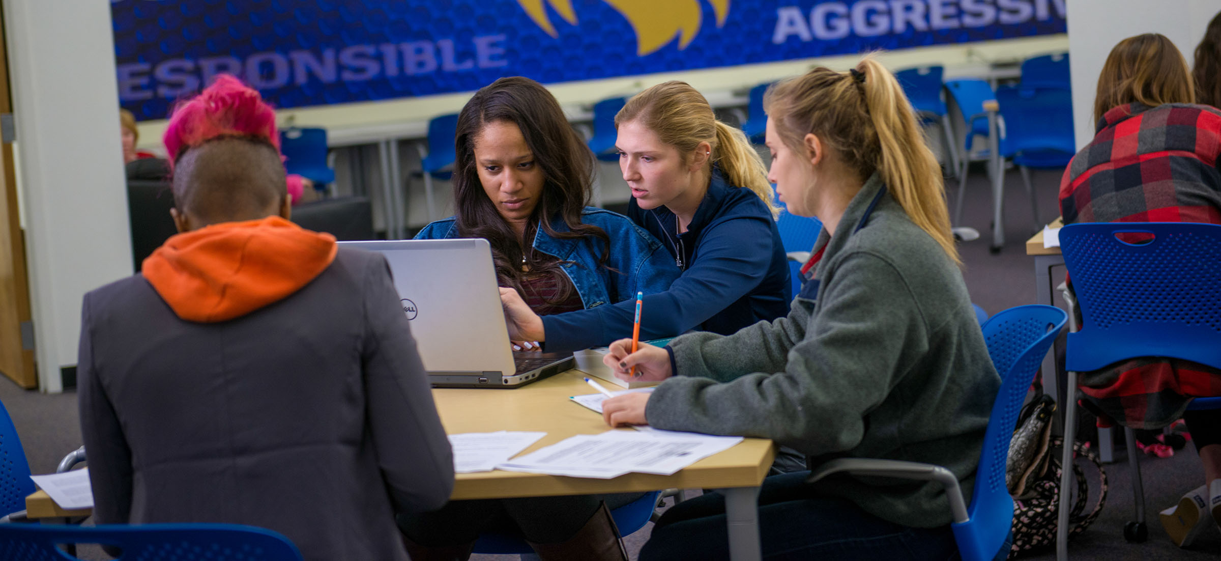A group of student completing an assignment together. They are at a desk with other desks around them. In the background there is blue with words on it.