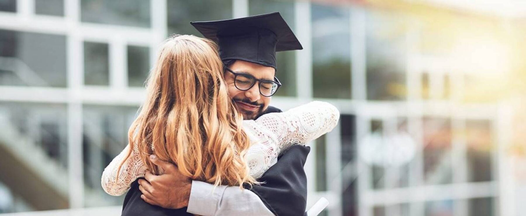A graduate in a cap and gown happily embraces a woman with long blonde hair. They are outside, in front of a modern building with glass windows. The graduate holds a diploma in one hand, and appears joyful and proud. Sunlight shines gently in the background.