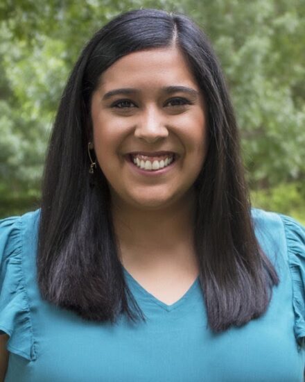 A smiling female with tanned skin and long dark brown hair. She is wearing a blue v neck blouse. In the background is trees.