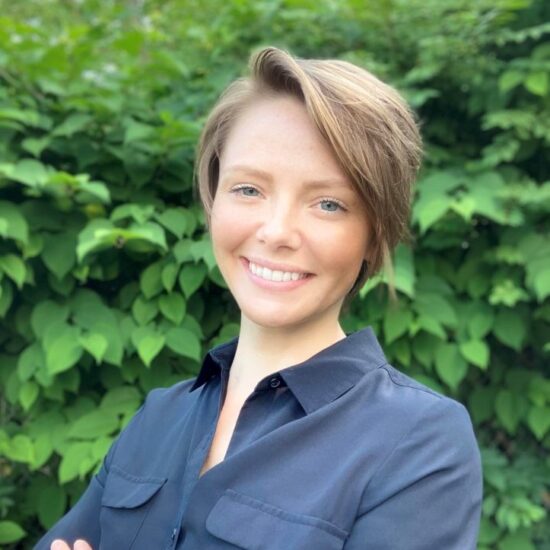 A smiling short hair dirty blonde female. She is wearing a black button up blouse. In the background is green trees.