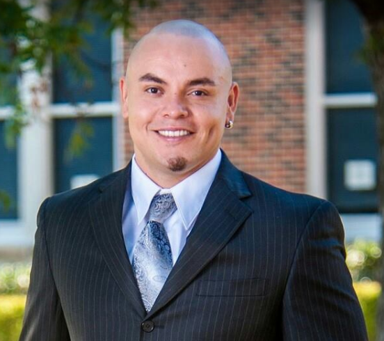 A bald young man smiling. He is wearing a black dress suit with a blue tie. In the background is a brick house with windows.