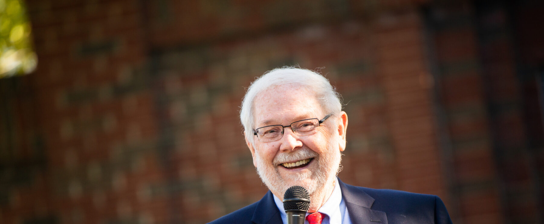 An elderly man with white hair and a beard is smiling and holding a microphone. He is wearing a dark blue suit, a light blue shirt, and a red tie. The background is a blurred view of a brick wall.