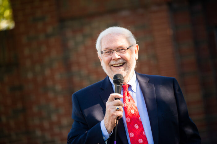 An elderly man with white hair and a beard is smiling and holding a microphone. He is wearing a dark blue suit, a light blue shirt, and a red tie. The background is a blurred view of a brick wall.