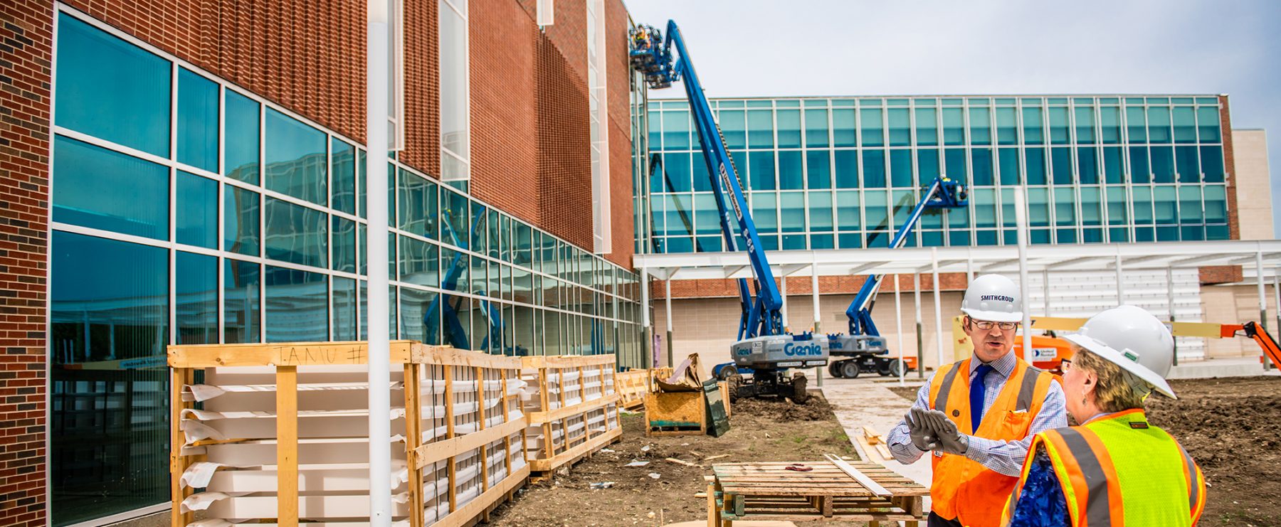Two construction workers wearing hard hats and orange safety vests discuss plans at a construction site. One worker gestures with their hands towards a partly constructed building.