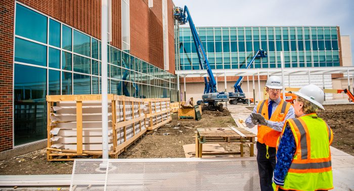 Two construction workers wearing hard hats and orange safety vests discuss plans at a construction site. One worker gestures with their hands towards a partly constructed building.