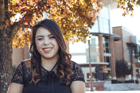 A young woman smiling with brown curled hair. She wearing a black short sleeve blouse. She is standing by a tree with autumn colored leaves. In the background there is a building with a lot of windows.