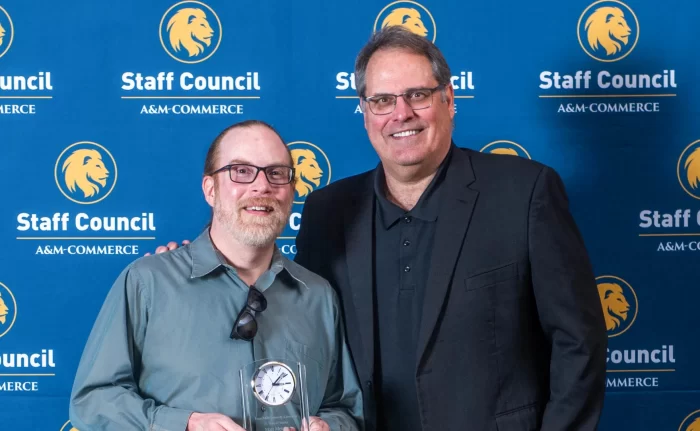 Two individuals are standing in front of a blue backdrop with the "Staff Council A&M-Commerce" logo. The person on the left is holding a glass award and smiling, while the person on the right is also smiling and has one arm around the other person.