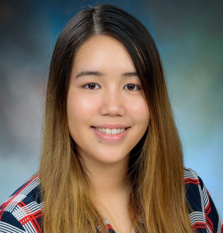 A young woman with long brown hair, wearing a plaid-patterned blouse, smiles for a headshot against a softly blurred blue and green background.