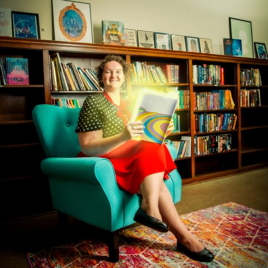 Dr. Rebecca Rowe seated in a chair with a book that has a rainbow coming out of it.