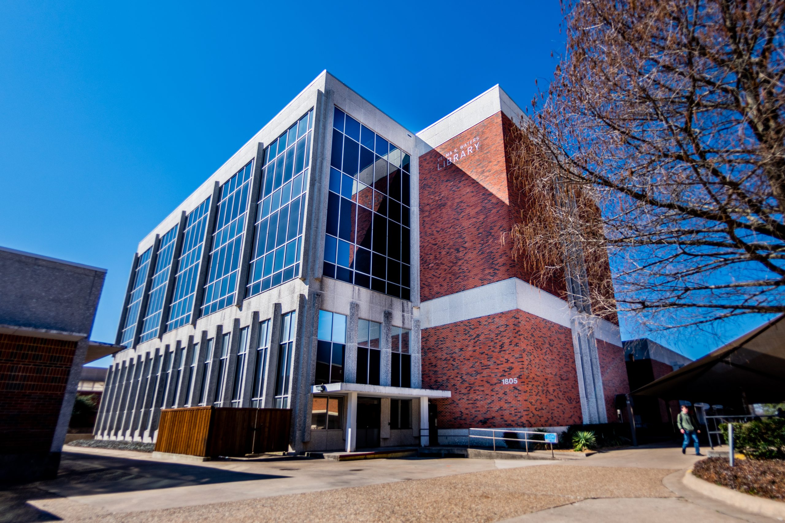 Modern university library building with glass facade and brick exterior. Clear blue sky background. Sign reads "Library" on the side