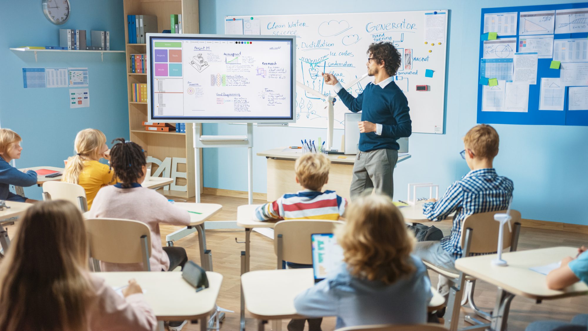 Man pointing to a white board in front of elementary students in desks