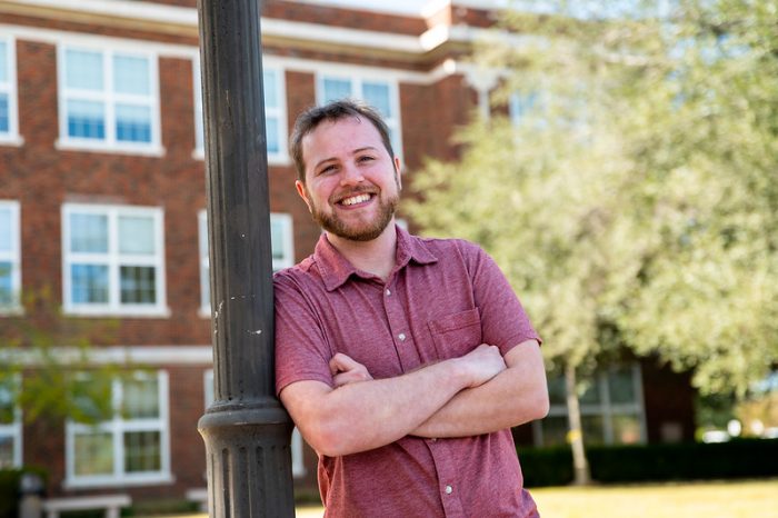 A smiling man in a short-sleeved red shirt stands outdoors, leaning against a black lamppost with arms crossed. A brick building with large windows is in the background, along with trees and greenery.