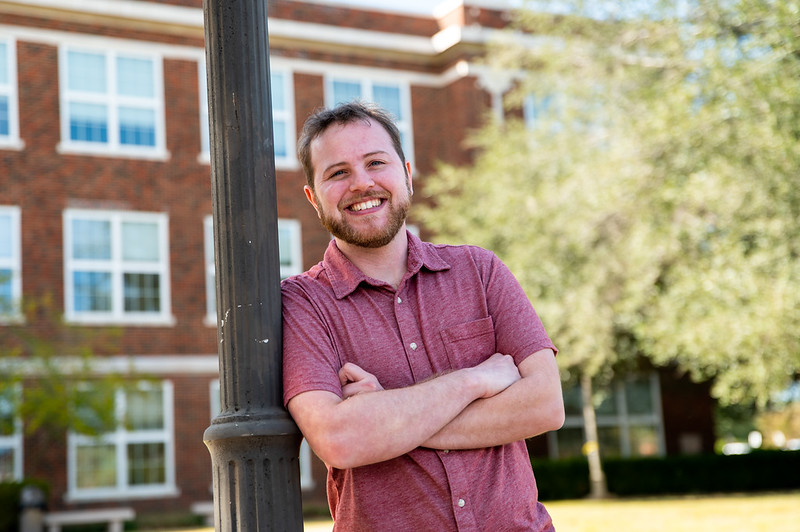 A smiling man in a short-sleeved red shirt stands outdoors, leaning against a black lamppost with arms crossed. A brick building with large windows is in the background, along with trees and greenery.