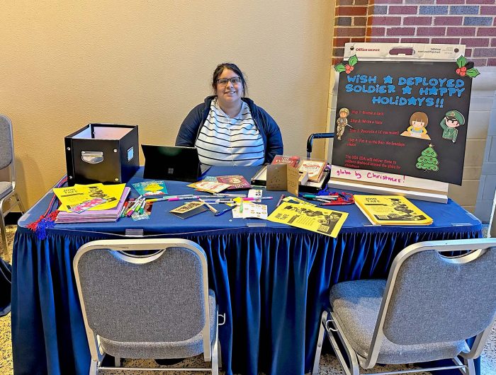 A person sits behind a table filled with colorful papers, markers, and brochures. A sign on the right reads, "Wish a deployed soldier a happy holiday!" The table has a blue tablecloth and two empty chairs in front. The setting appears to be an indoor event.