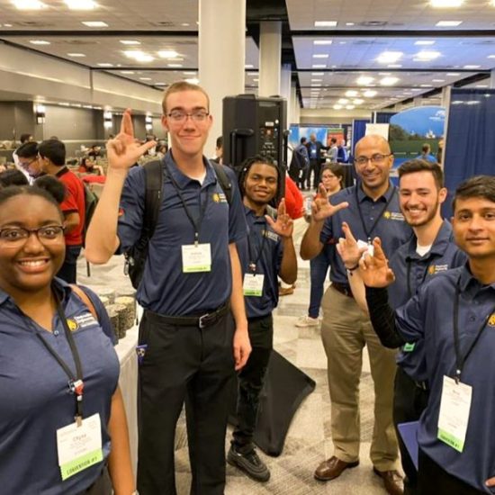 A group of six people, standing in a convention center wearing matching blue polo shirts. They are smiling and making hand gestures. The background features booths and other people mingling.