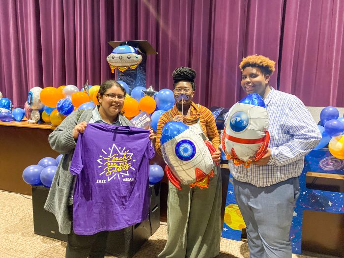 Three people are posing in front of a colorful backdrop with balloons. One person on the left is holding a purple T-shirt that says "Reach for the Stars 2022." The other two are each holding balloons shaped like rockets.