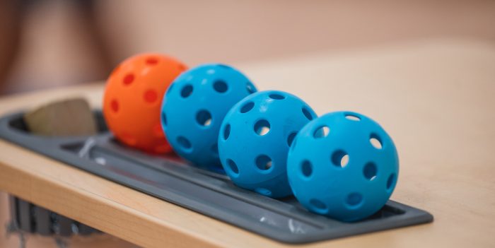 Four perforated plastic balls, two blue and two orange, are lined up in an orderly fashion on a beige tabletop. The balls rest in a grey tray, and their design suggests they are for a sport or game such as pickleball. The background is blurred.