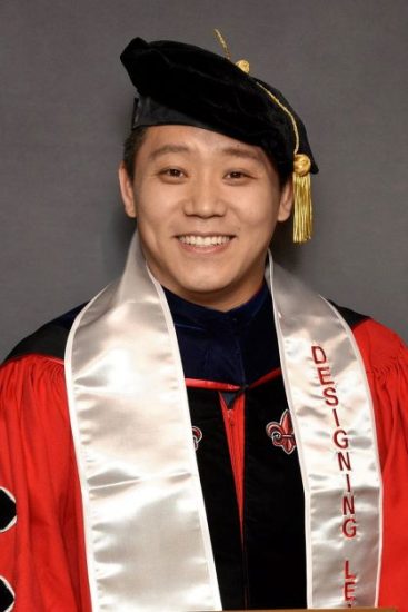A graduate dressed in an academic gown and cap smiles warmly at the camera. He wears a black academic hat and a white stole with red lettering that reads "Designing Leaders.