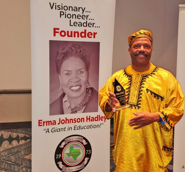 A man dressed in a bright yellow traditional African outfit holds an award while standing next to a banner. The banner features a large portrait of Erma Johnson Hadley with the words above her image and title below.