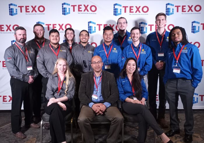 A group of thirteen individuals poses for a photo at a TEXO event. Most are wearing branded shirts in gray or blue. A background banner features the TEXO logo, which includes a blue building block graphic and red text. They all wear name badges.