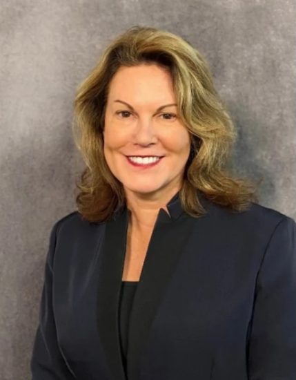 A smiling woman with shoulder-length, wavy blond hair wearing a navy blazer over a black top stands against a textured gray background. She appears confident and professional.