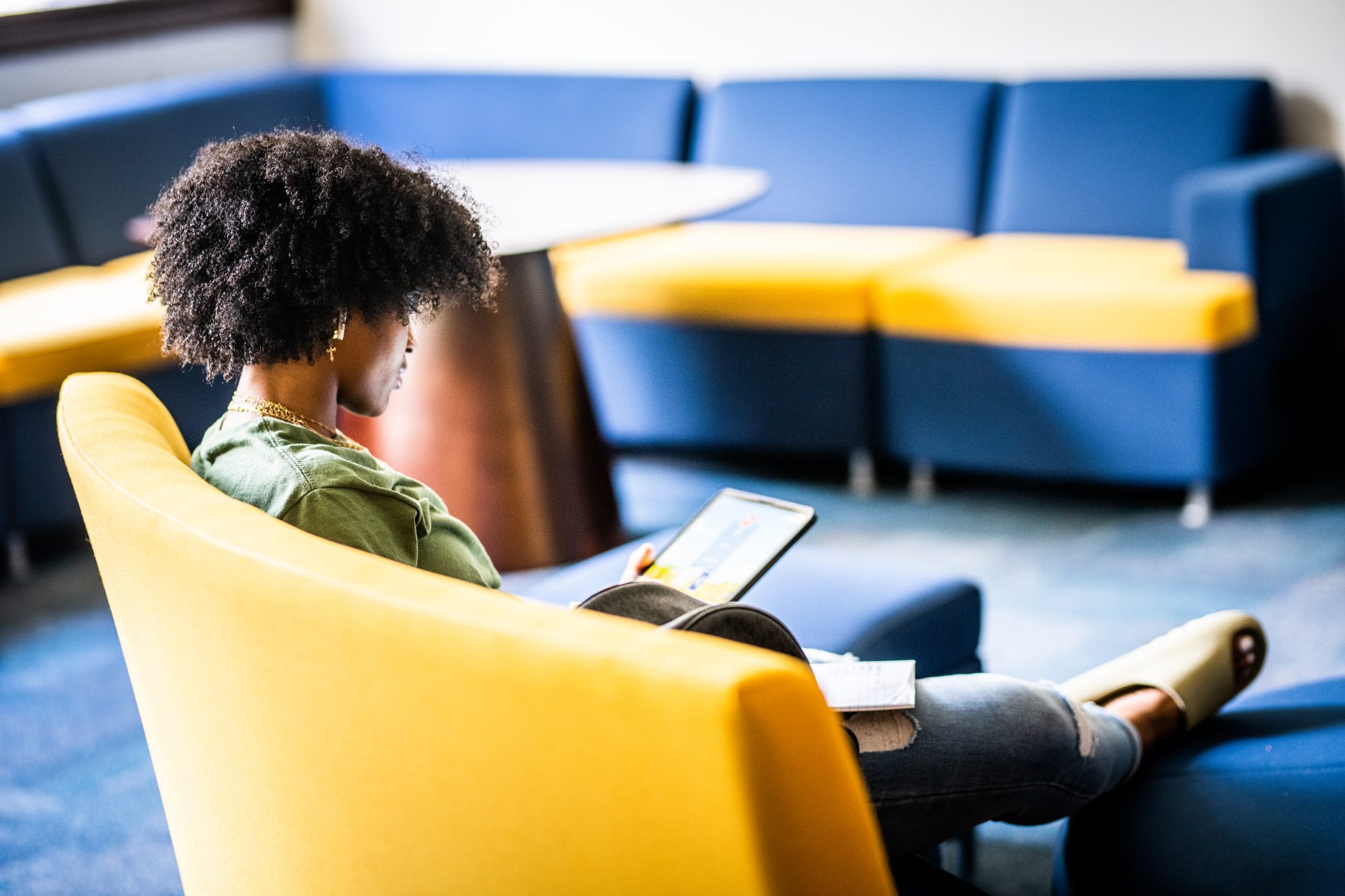 A student sits in a lounge area with a yellow and blue color scheme, focused on a tablet in their lap.