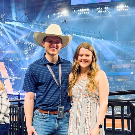 A man and a woman smile while posing for a picture in an indoor arena. The man wears a cowboy hat, blue polo shirt, and jeans. The woman wears a striped dress. Bright stage lights and a large structure are visible in the background.